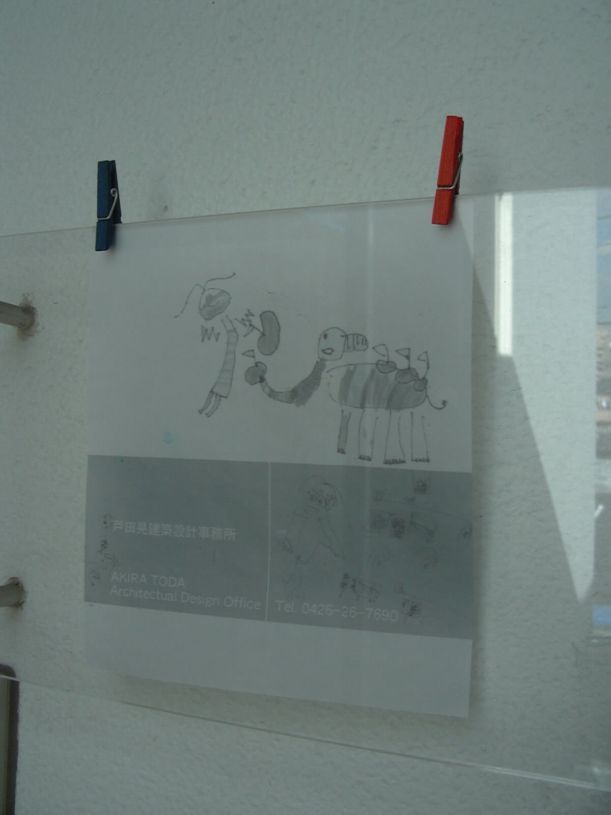 透明の板に洗濯バサミで取り付けられた、白い紙に子供のイラストと電話番号が書かれている戸田晃建築設計事務所の看板